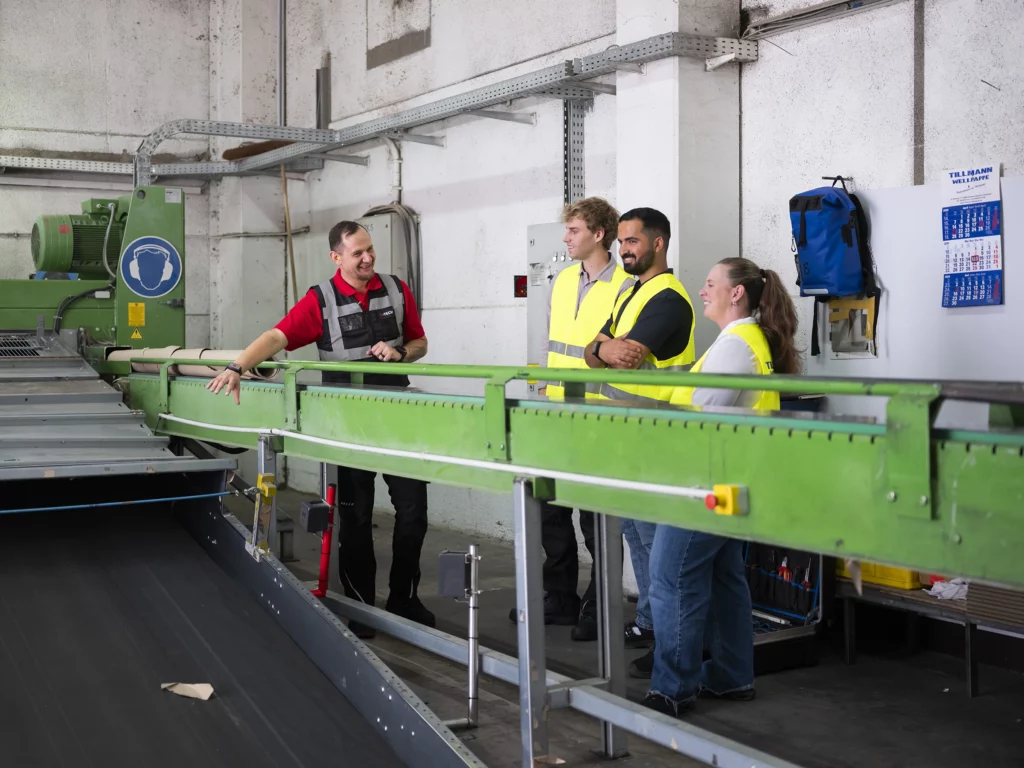 Personen in Sicherheitswesten neben einer grünen Maschine in einer Fabrikhalle.