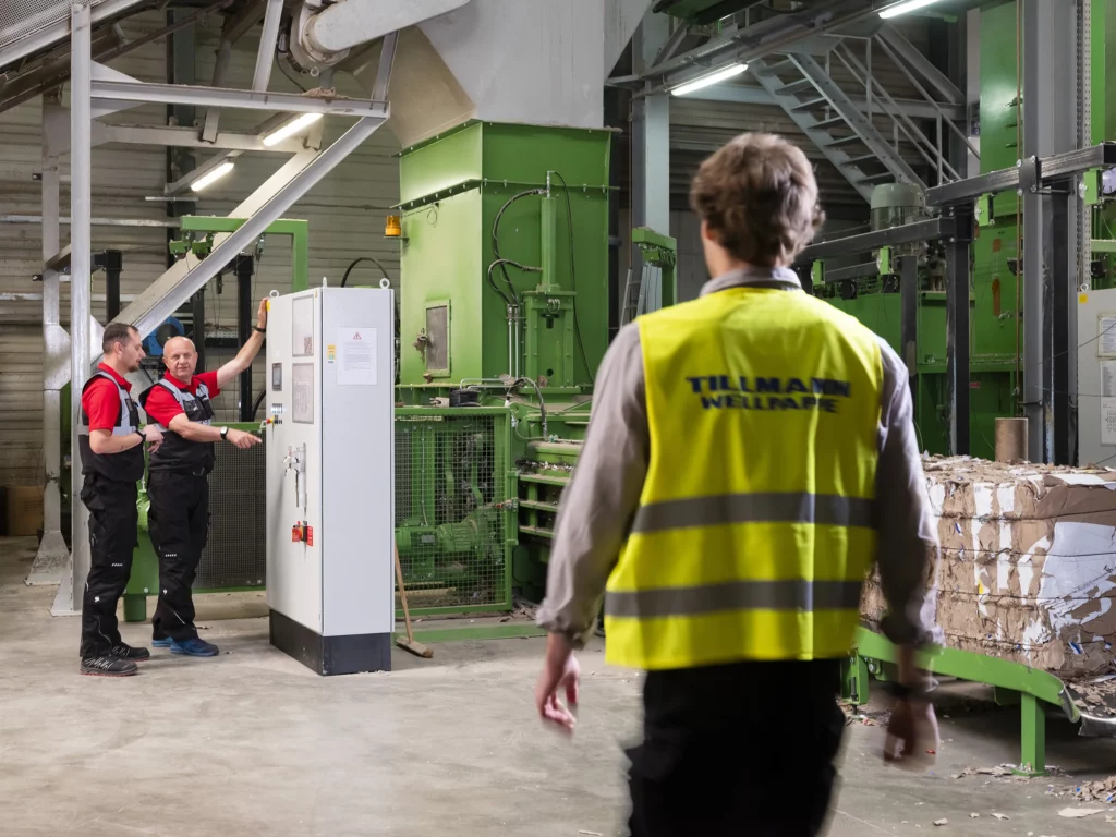 Drei Männer in Fabrik, einer mit gelber Weste, zwei an Schaltschrank.